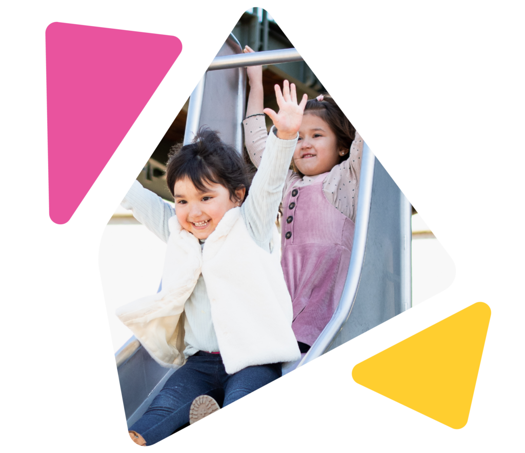 two young girls smiling, sliding down outdoor slide with arms up