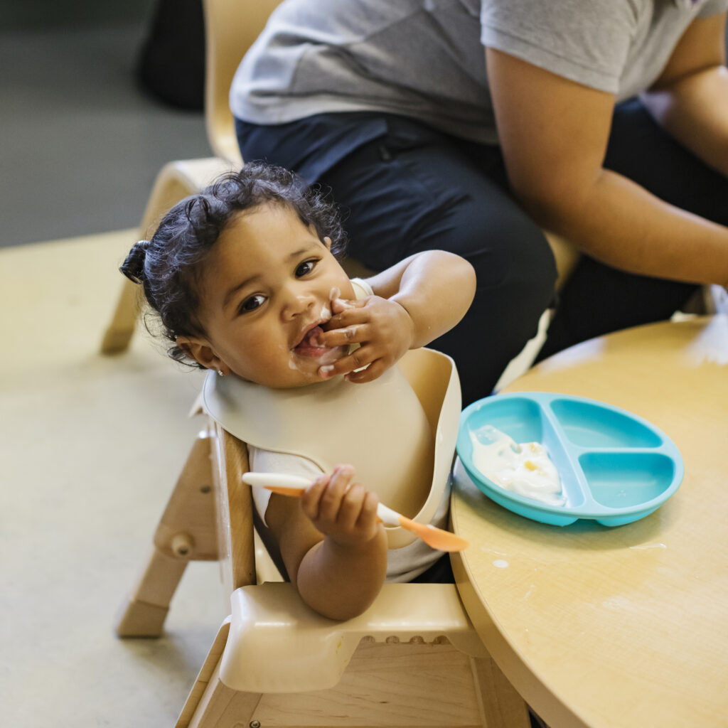 A toddler sits at a table in a daycare, feeding herself